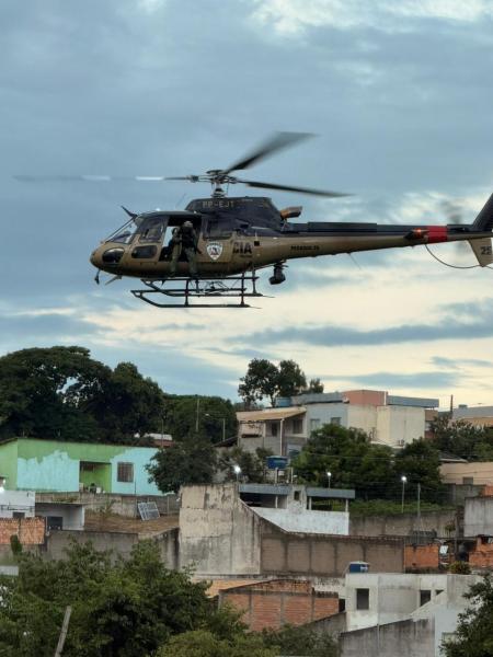 Helicóptero da polícia sobrevoando uma área urbana com casas e vegetação ao fundo.