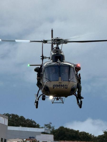 Helicóptero da PMMG voando em um céu nublado, com luzes verdes e vermelhas acesas.
