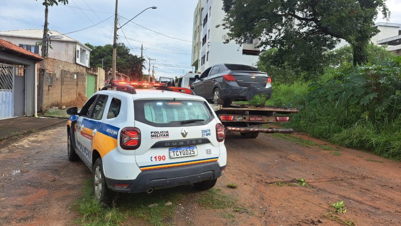 Viatura da Polícia Militar parada em uma rua de terra, com um carro sendo rebocado por uma caminhonete ao fundo.
