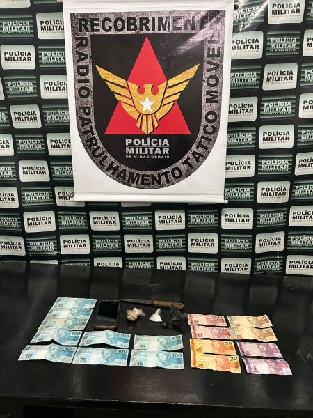 Imagem de apreensão policial com cédulas de dinheiro em diferentes denominações e objetos suspeitos dispostos sobre uma mesa. Ao fundo, um banner da Polícia Militar com o emblema da corporação.
