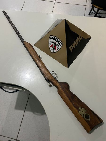 Rifle com corpo de madeira posicionado ao lado de um objeto em forma de pirâmide com o logo PMMG.