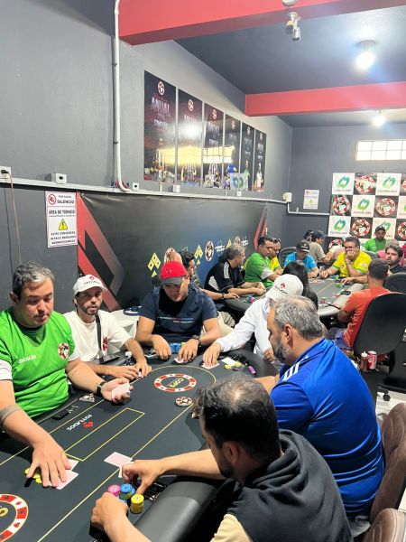 A lively poker room filled with players sitting around tables, engaged in a game. Participants are diverse, wearing casual clothing and engaging with poker chips and cards. The walls feature gaming-related artwork and signage.