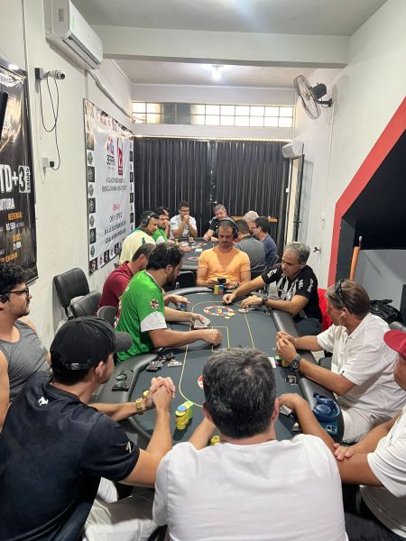 A group of people engaged in a poker game at a table, showing multiple players with chips and cards in a casual indoor setting.