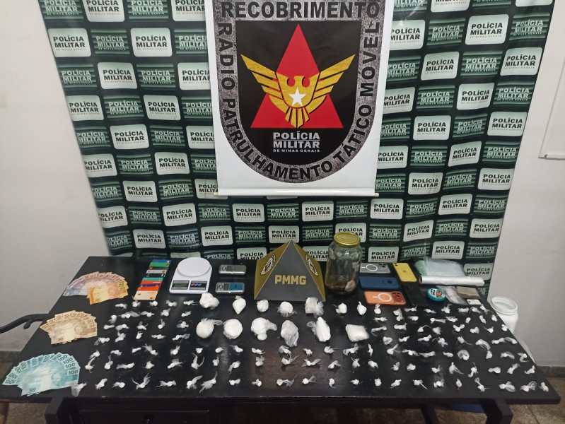 Mesa com drogas, balanças, maços de dinheiro e outros itens apreendidos pela polícia, com fundo de painel da Polícia Militar.