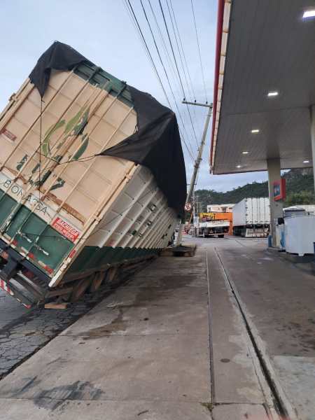 Caminhão tombado em um posto de gasolina, com a carroceria inclinada e coberta por uma lona preta.
