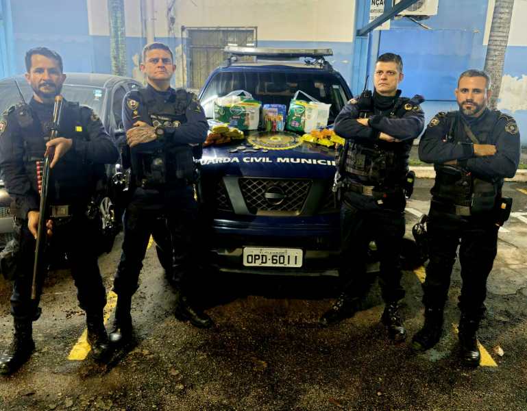 Quatro oficiais da Guarda Civil Municipal em frente a uma viatura, com itens de doação sobre o capô.