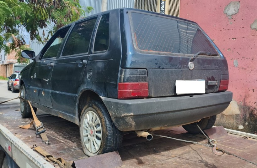 Um carro preto, modelo hatchback, estacionado em uma plataforma de reboque, com detalhes sujos e arranhões visíveis. A parte traseira e lateral do veículo estão visíveis, com rodas de liga leve e um escapamento visível.
