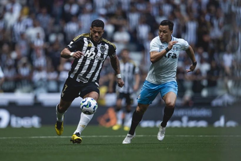 A soccer game action shot featuring two players in motion, one in black and white stripes and the other in light blue, competing for the ball on the field.