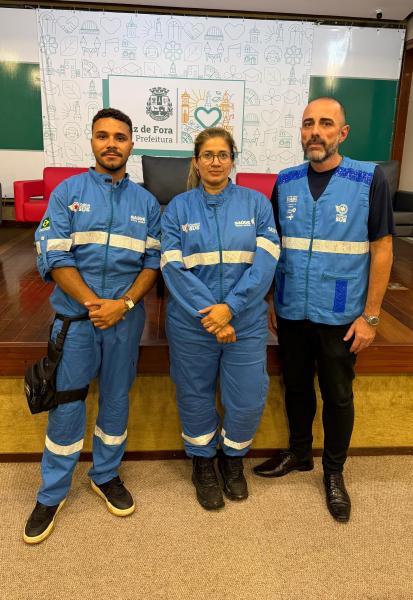 Três pessoas vestindo uniformes, duas em macacões azuis e uma em colete azul, posando em um cenário institucional com o logotipo da Prefeitura de Juiz de Fora ao fundo.