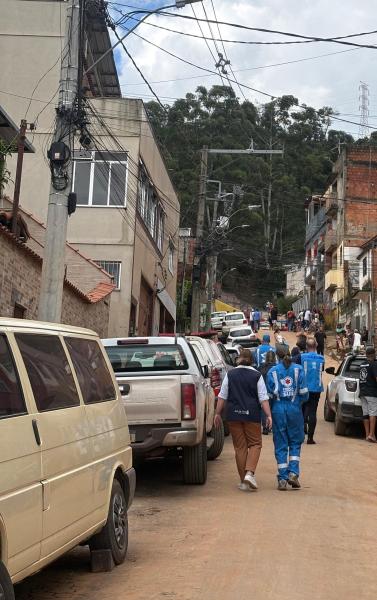 Caminho em uma rua de moradia com várias casas e veículos estacionados, onde pessoas com uniformes azuis caminham em direção à colina.
