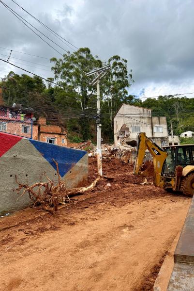 Vista de uma área afetada por deslizamento de terra, com um maquinário de construção ao lado de entulho e árvores caídas, sob um céu nublado.