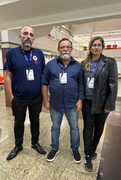 Três pessoas posando para a foto em um ambiente interno, vestindo roupas casuais e crachás de identificação.