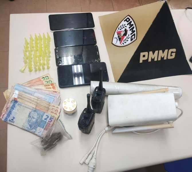 Imagem de objetos apreendidos, incluindo telefones celulares, dinheiro em notas de diferentes valores, material suspeito em formato de pacotes, rádios comunicadores e uma placa com o logo da PMMG.