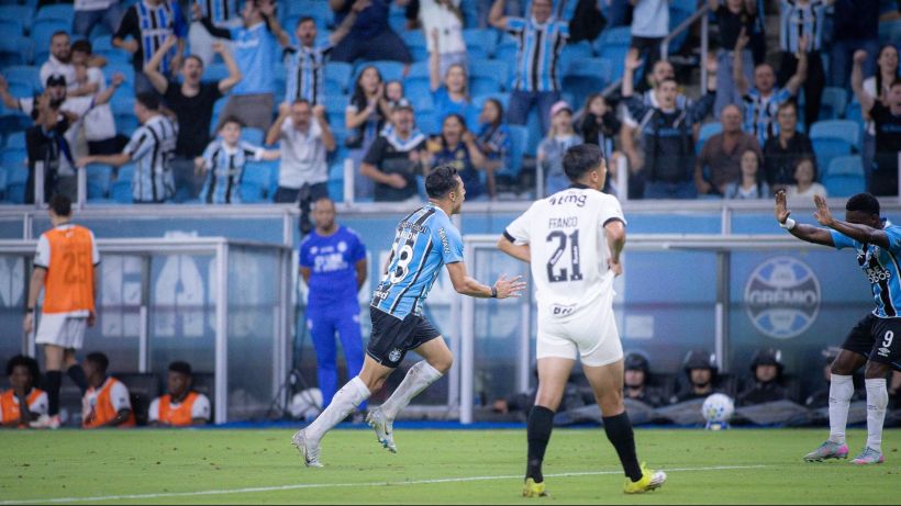 A soccer player in a blue and black uniform celebrates after scoring a goal, while fans cheer in the background. Opposing players in white uniforms look on.