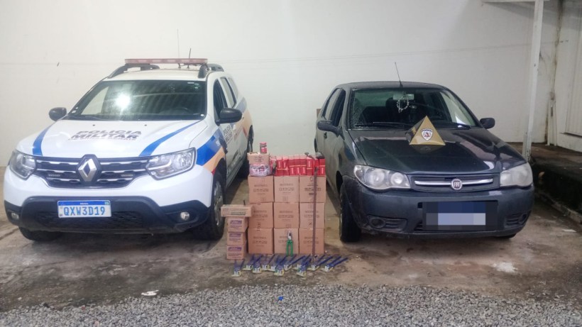 Foto de um carro da polícia militar ao lado de um veículo particular. No chão, há várias caixas e objetos, possivelmente evidências de uma apreensão.