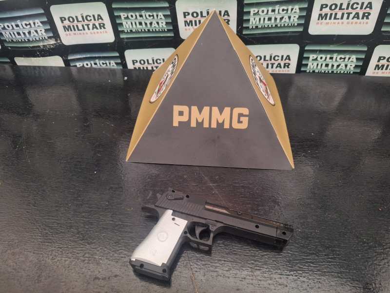 Imagem de uma pistola semi-automática ao lado de uma pirâmide com a sigla PMMG, em um fundo com placas da Polícia Militar.