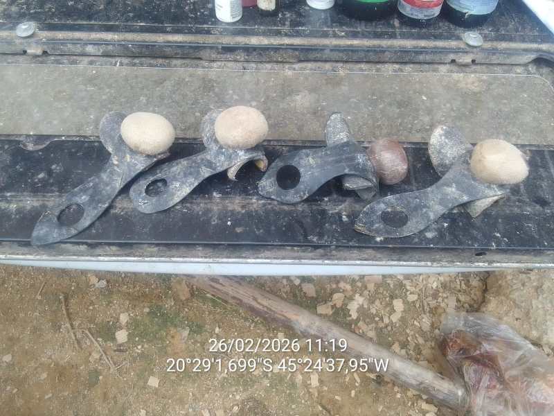 Quatro suportes de metal com pedras na parte superior, dispostos em uma superfície preta, com fundo de terra.