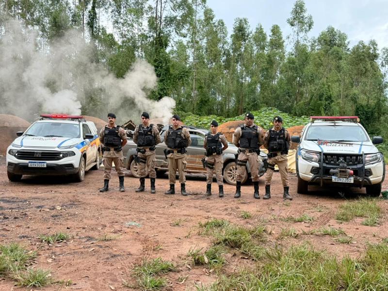 Grupo de policiais em uniforme tático posando em uma área rural, com veículos de polícia ao fundo e fumaça saindo de um local próximo.