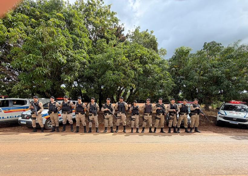 Grupo de policiais uniformizados em pé ao lado de viaturas, com árvores ao fundo.