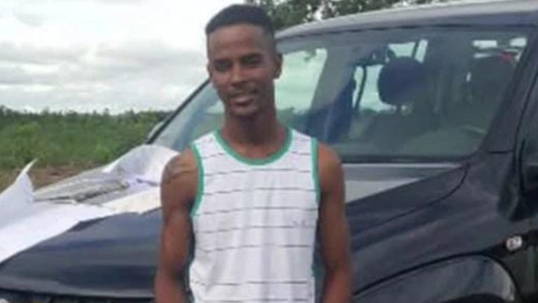 Homem em camiseta listrada posando ao lado de um veículo em uma área rural.