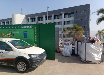 Veículo estacionado em frente a um portão verde, com uma construção ao fundo. Há materiais recicláveis acumulados ao lado do portão.