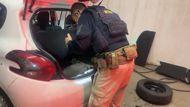 Policial inspecionando o porta-malas de um carro branco durante uma operação.