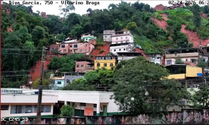 Vista de uma comunidade em um morro com casas coloridas, vegetação densa e fios elétricos ao fundo.