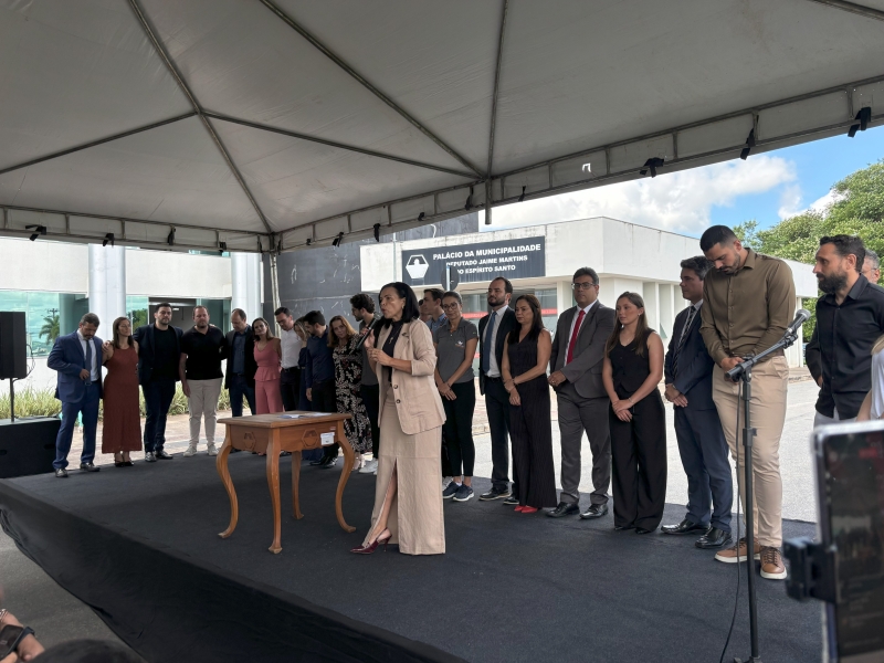 Evento ao ar livre com um grupo de pessoas em um palco, onde uma mulher no centro fala ao microfone. Elas estão posicionadas em frente a um cartaz do 'Palácio da Municipalidade'.