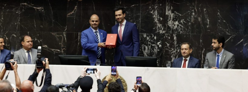 Cerimônia formal com políticos apresentando um documento, enquanto outros observam e registram com celulares.