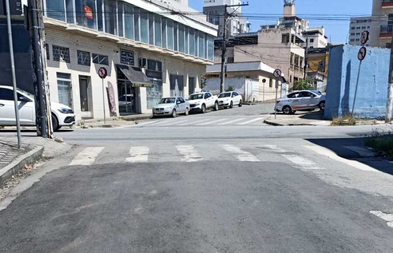 Vista de uma interseção de ruas, com calçada e sinalizações de trânsito visíveis. Vários carros estão estacionados ao longo da rua e prédios comerciais estão ao fundo.