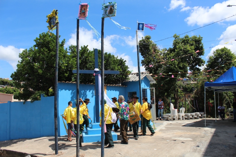 Grupo de pessoas vestindo camisetas amarelas se reunindo em um espaço ao ar livre com uma casa azul ao fundo, decorada com bandeirinhas coloridas. No centro, uma cruz e bandeirinhas penduradas em postes.