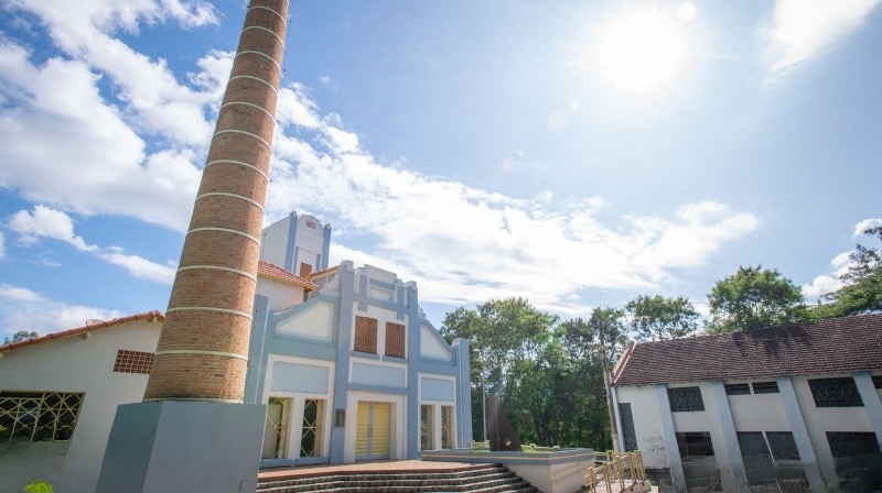 Prédio histórico com chaminé ao fundo, céu azul e árvores ao redor.