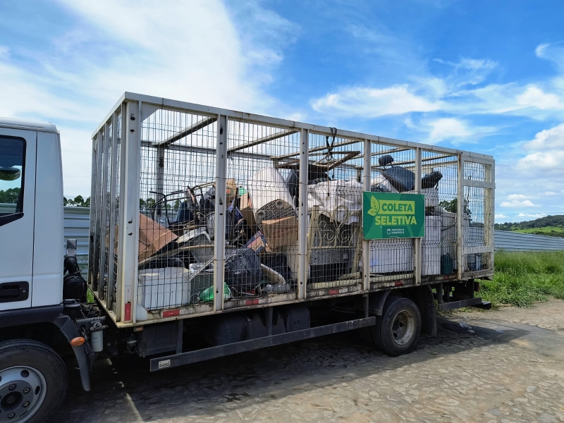Caminhão de coleta seletiva com carga de materiais recicláveis, incluindo móveis e resíduos, estacionado sob um céu azul claro.