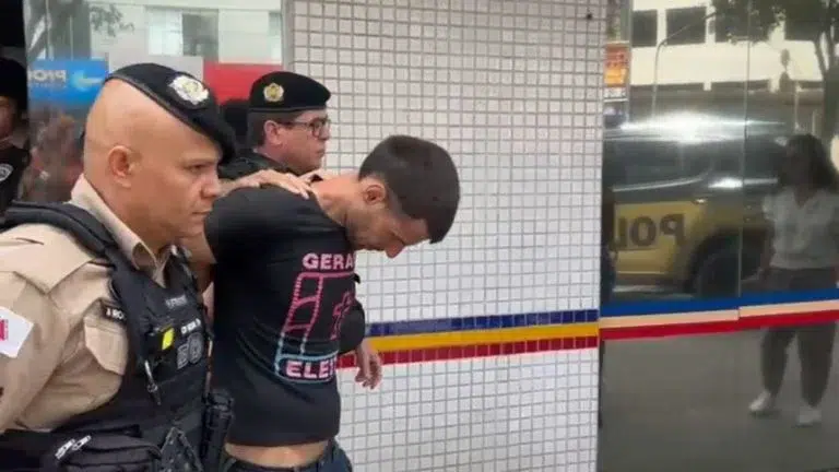 Um homem sendo levado por policiais em uma abordagem na rua, com uma parede azulejada ao fundo.