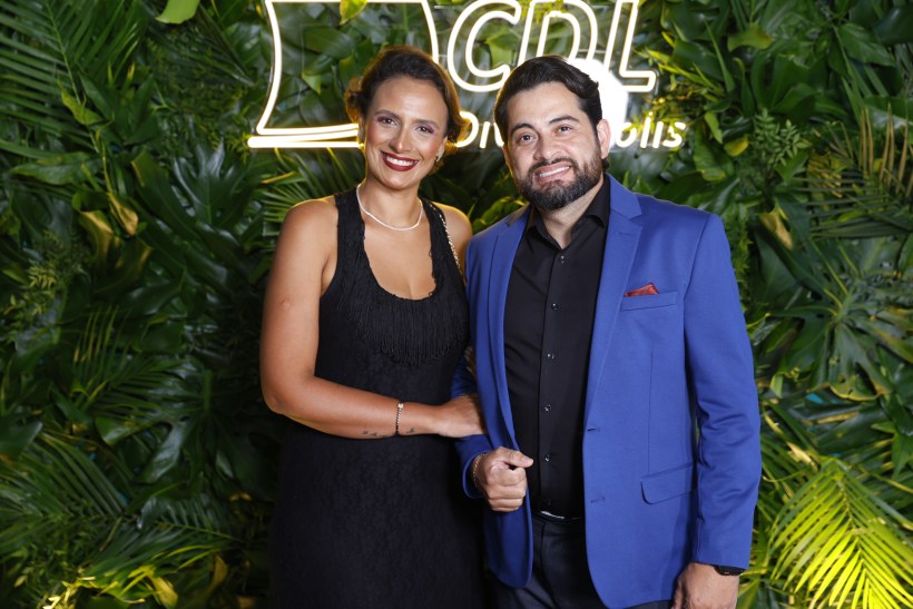 Casal posando em evento, com fundo de folhagens verdes e iluminação com o nome 'CDL'. A mulher veste um vestido preto com franjas e o homem usa um blazer azul.