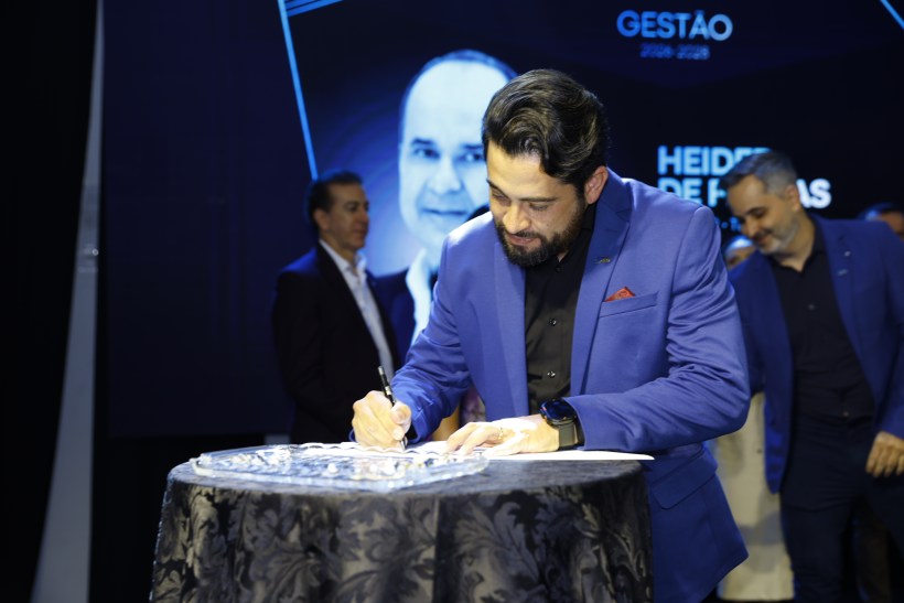 Homem assinando um documento em uma mesa durante um evento, com um fundo que exibe um retrato e o texto 'GESTÃO 2020-2023'.