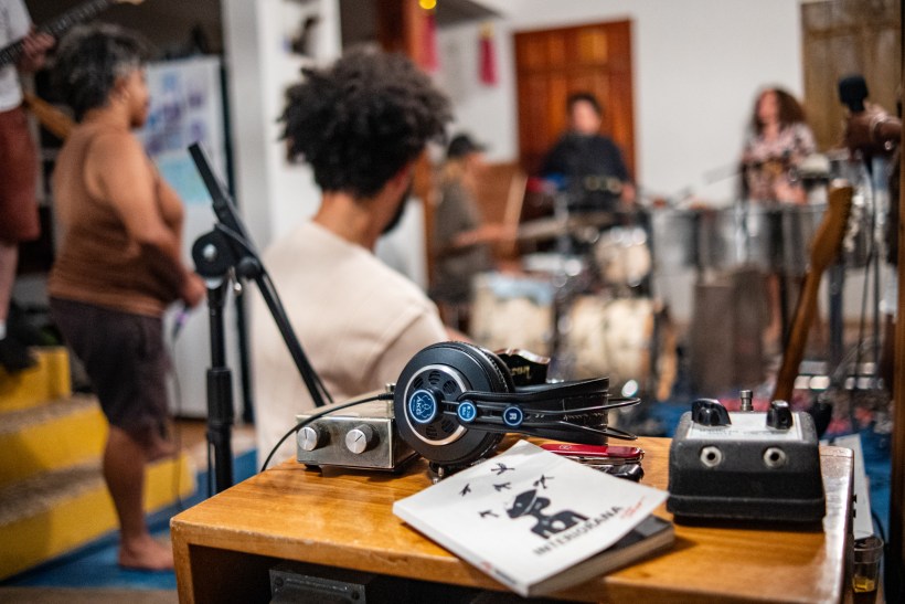Imagem de um ensaio musical, mostrando uma mesa com equipamentos de áudio, fones de ouvido e um caderno em primeiro plano, enquanto músicos se apresentam ao fundo.