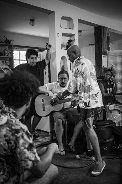 Grupo de pessoas em um ambiente interno, com um homem tocando violão enquanto outros observam e interagem, criando um clima de descontração e música.