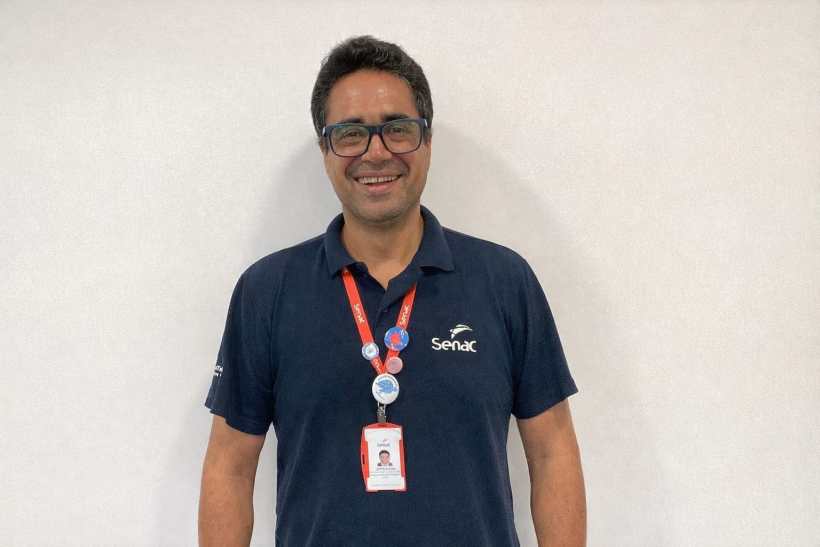 Homem sorridente usando camiseta azul escura com o logo do Senac, pendurado em um crachá e com bottons no colarinho.