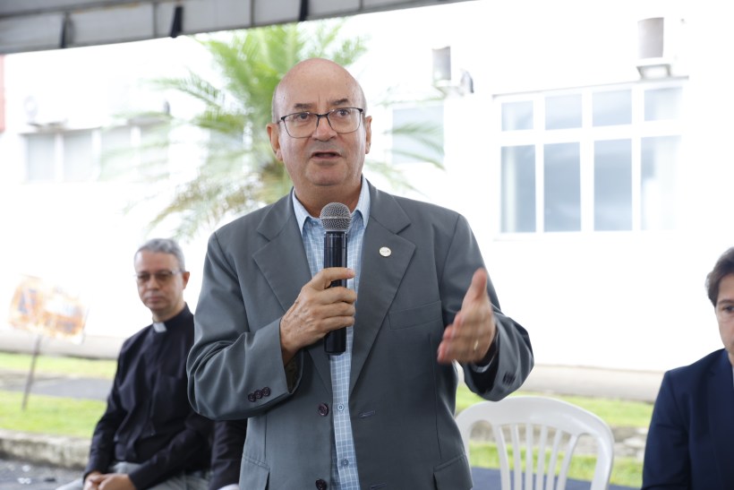 Homem de óculos e paletó cinza segurando um microfone e falando em um evento, com pessoas sentadas ao fundo.
