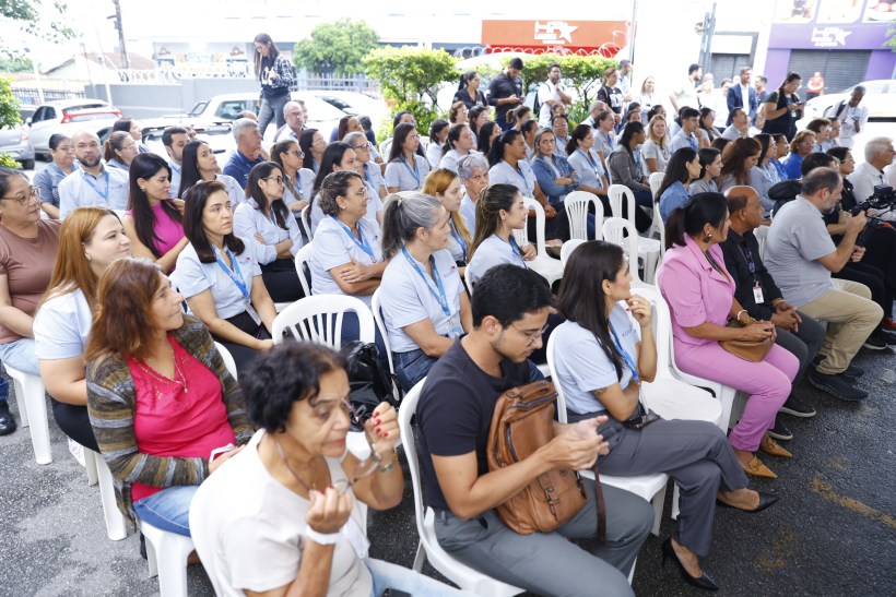 Grupo de pessoas sentadas em cadeiras plásticas, assistindo a um evento ao ar livre. Muitas delas vestem camisetas claras e há uma variedade de estilos de cabelo. Algumas pessoas interagem com dispositivos móveis.