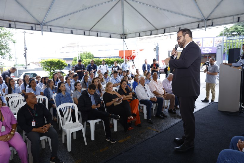 Evento ao ar livre com uma grande plateia sentada em cadeiras brancas, enquanto um palestrante se dirige ao público. O cenário inclui um toldo e uma faixa de vegetação ao fundo.