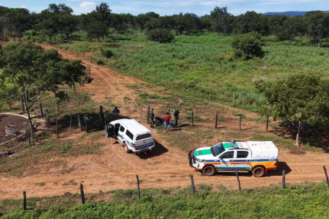 Vista aérea de um terreno rural com um caminho de terra, onde estão estacionadas uma van e uma viatura policial. Algumas pessoas estão reunidas próximo à viatura, cercadas por vegetação.