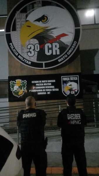 Dois policiais de costas em frente à fachada da Polícia Militar do Mato Grosso, com logotipos e faixas demonstrando suas respectivas unidades.
