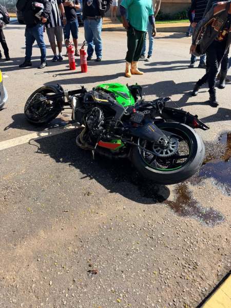 Motocicleta acidentada deitada no chão, com sujeira e óleo ao redor, enquanto pessoas observam ao fundo.