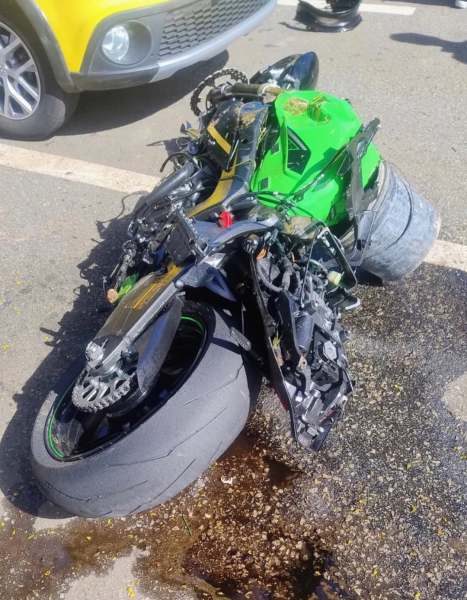 Motocicleta danificada de cor verde deitada no chão, com partes quebradas e óleo no asfalto ao redor.