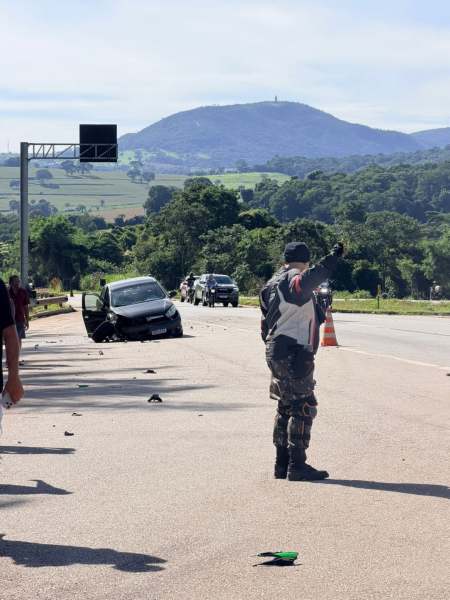 Um policial sinaliza para o tráfego em uma estrada, próximo a um carro acidentado, com montanhas ao fundo.