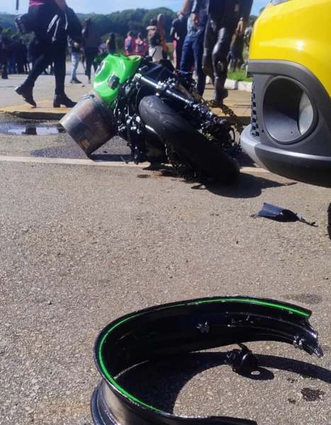Motocicleta verde caída no chão, com danos visíveis, ao lado de um carro amarelo e uma multidão ao fundo.