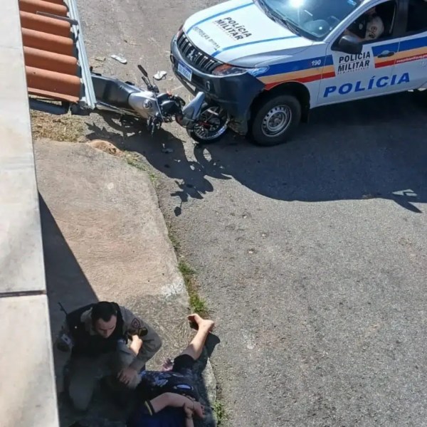 Um policial prestando atendimento a uma pessoa caída no chão, enquanto uma viatura da polícia militar está estacionada próximo a uma motocicleta acidentada.