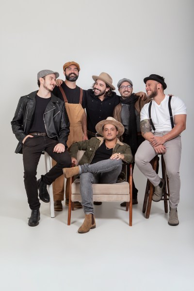Seis homens posando em estúdio, usando roupas casuais e chapéus, com um sentado em uma cadeira e os demais em pé ao redor.